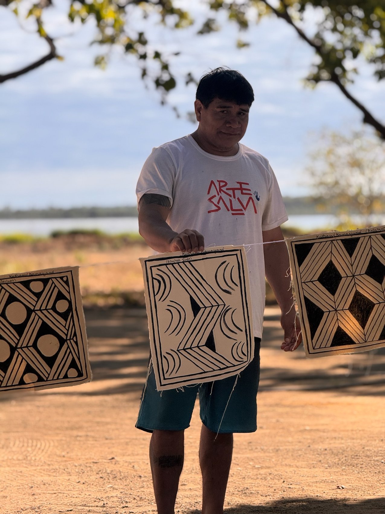 Kamayurá com camiseta Arte Salva segurando painéis de grafismo ao ar livre